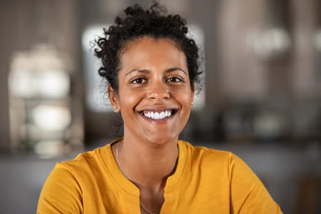 Smiling woman with curly hair in a mustard-yellow top, looking at the camera in a warmly lit room.