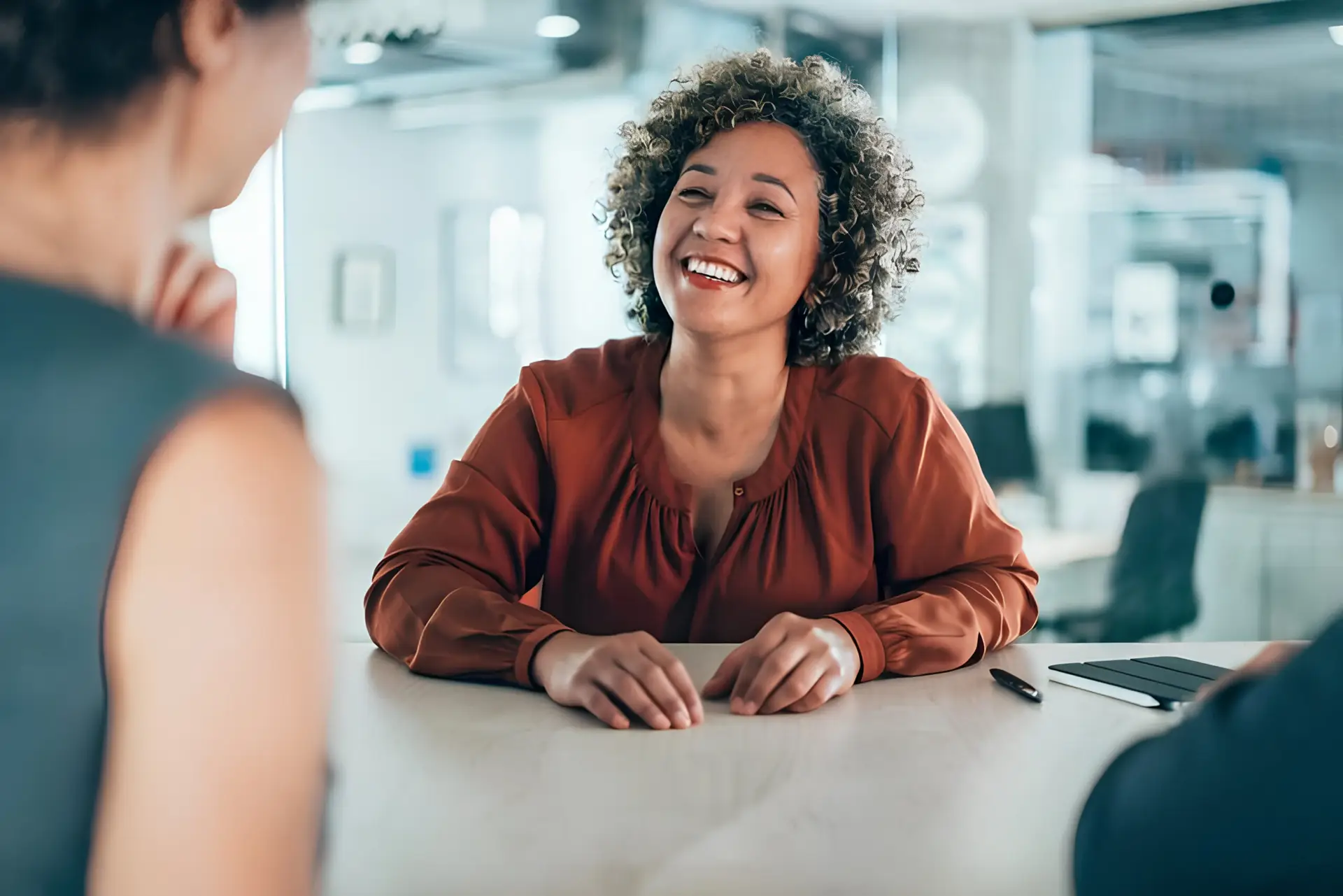 Smiling woman with curly hair in a rust-colored blouse seated at a white table, talking with a colleague in a bright office.