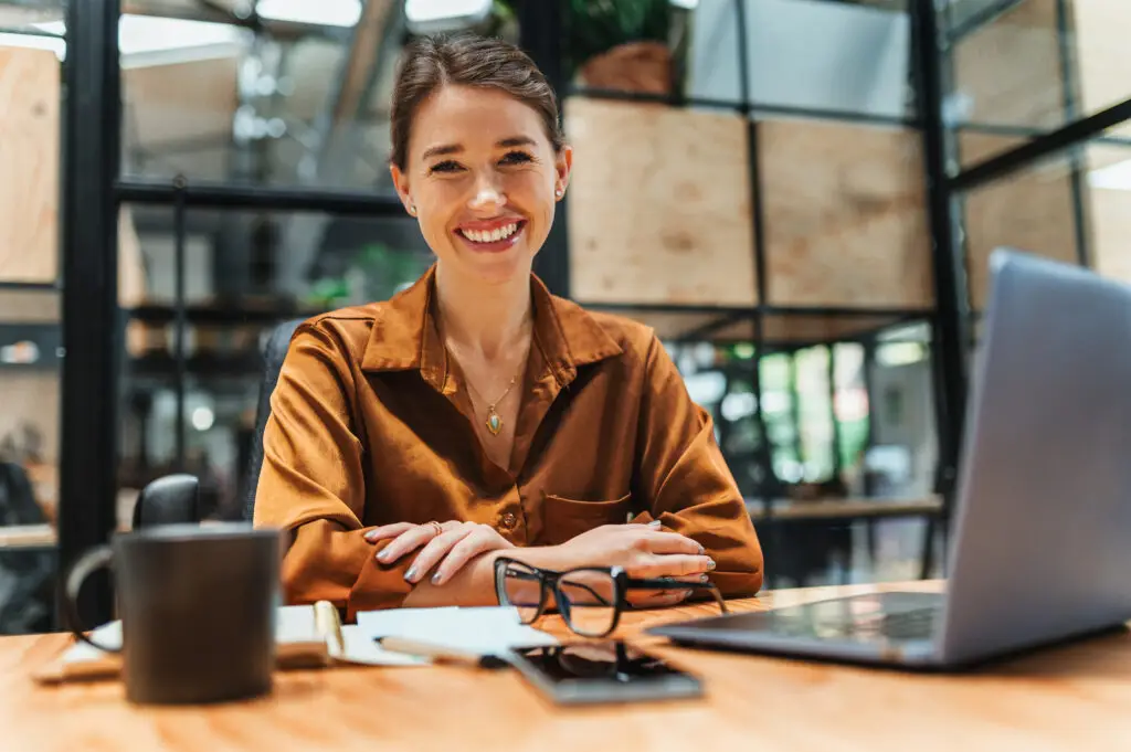 Professional coach in a rust silk top smiling at a desk with a laptop, notebook, and glasses.