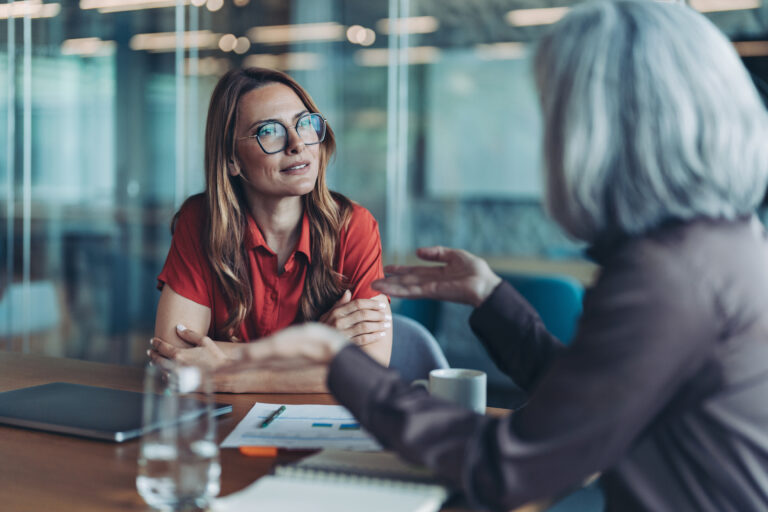 Two colleagues in a one-on-one conversation at a meeting table in a glass-walled office.