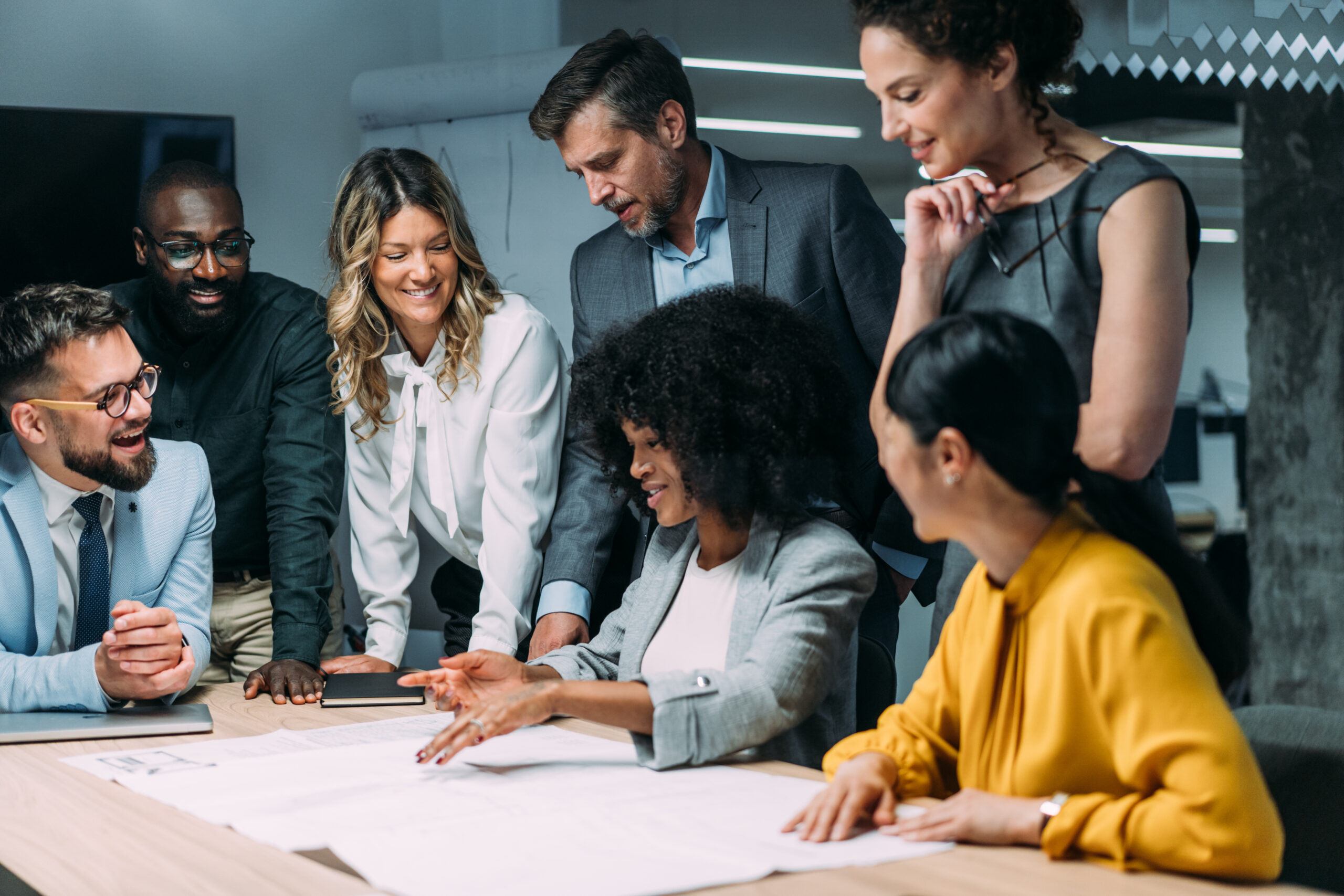 Seven colleagues around a conference table looking at a large document spread in front of them, one woman gesturing toward it as the others watch