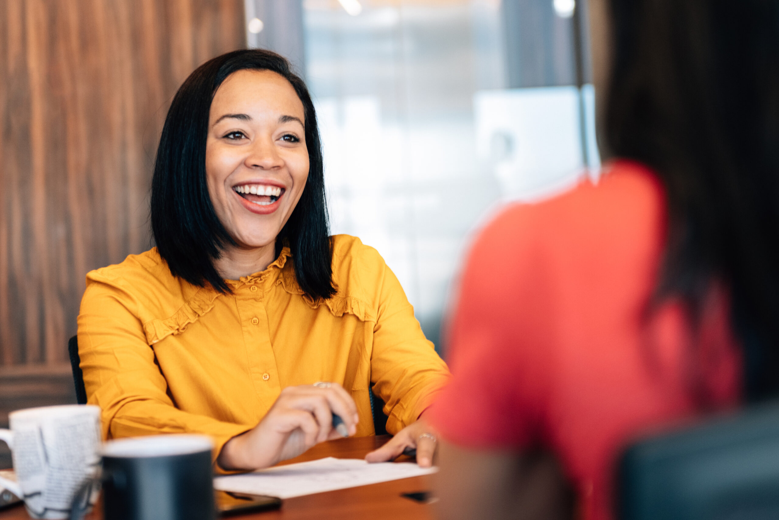 Woman in a yellow blouse laughing during a one-to-one conversation across a table