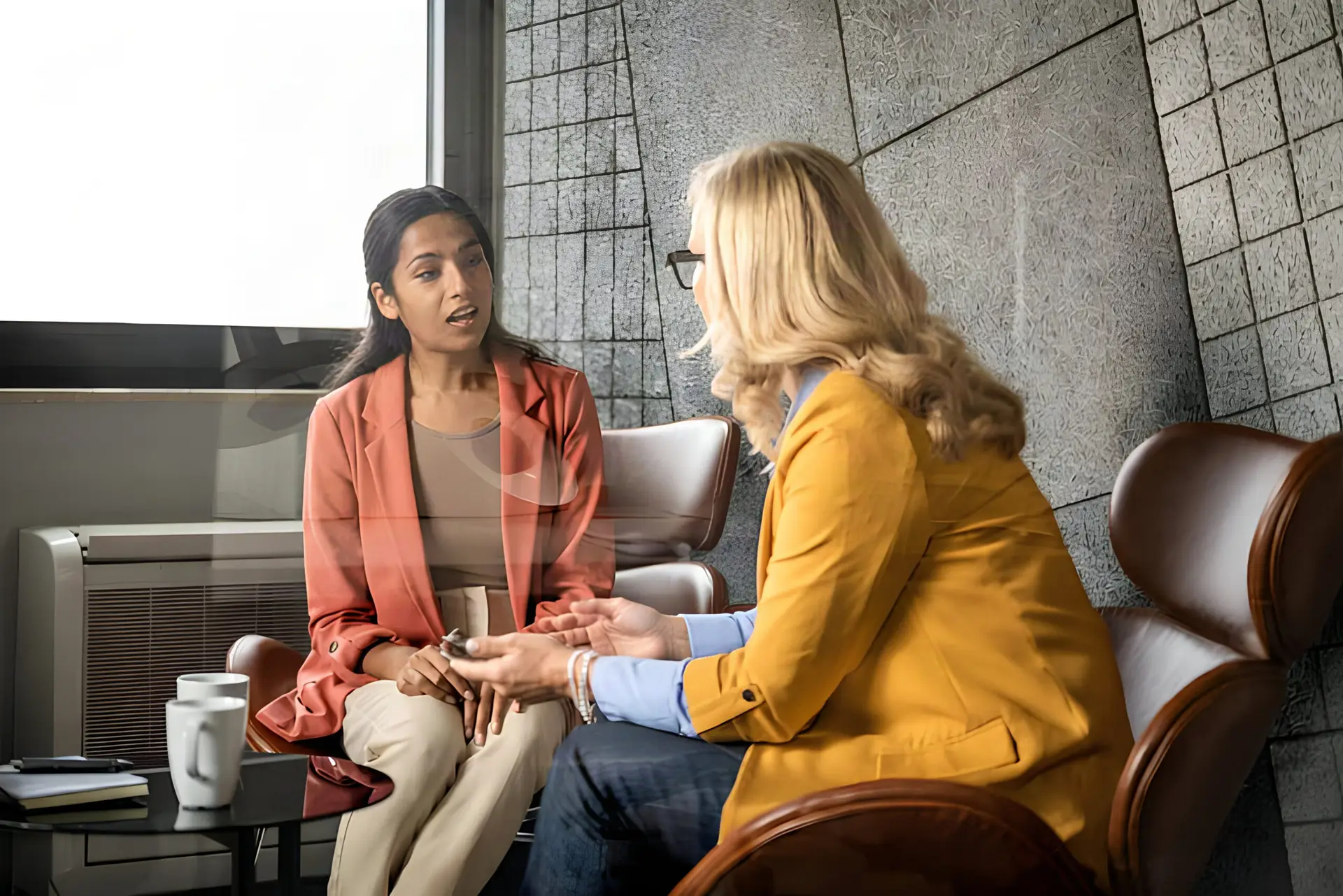Two women seated in armchairs in conversation, one listening while the other speaks