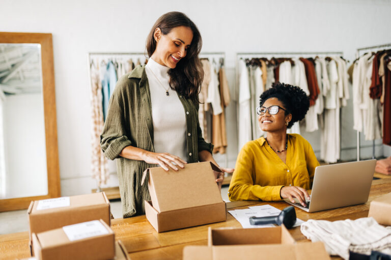 Two women in a clothing store, one packing a cardboard box and the other working at a laptop, with shipping boxes and stock around them