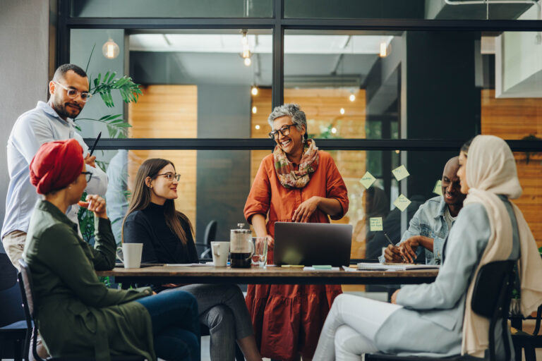 Group of colleagues in an open meeting space, talking around a table with laptops and notes.