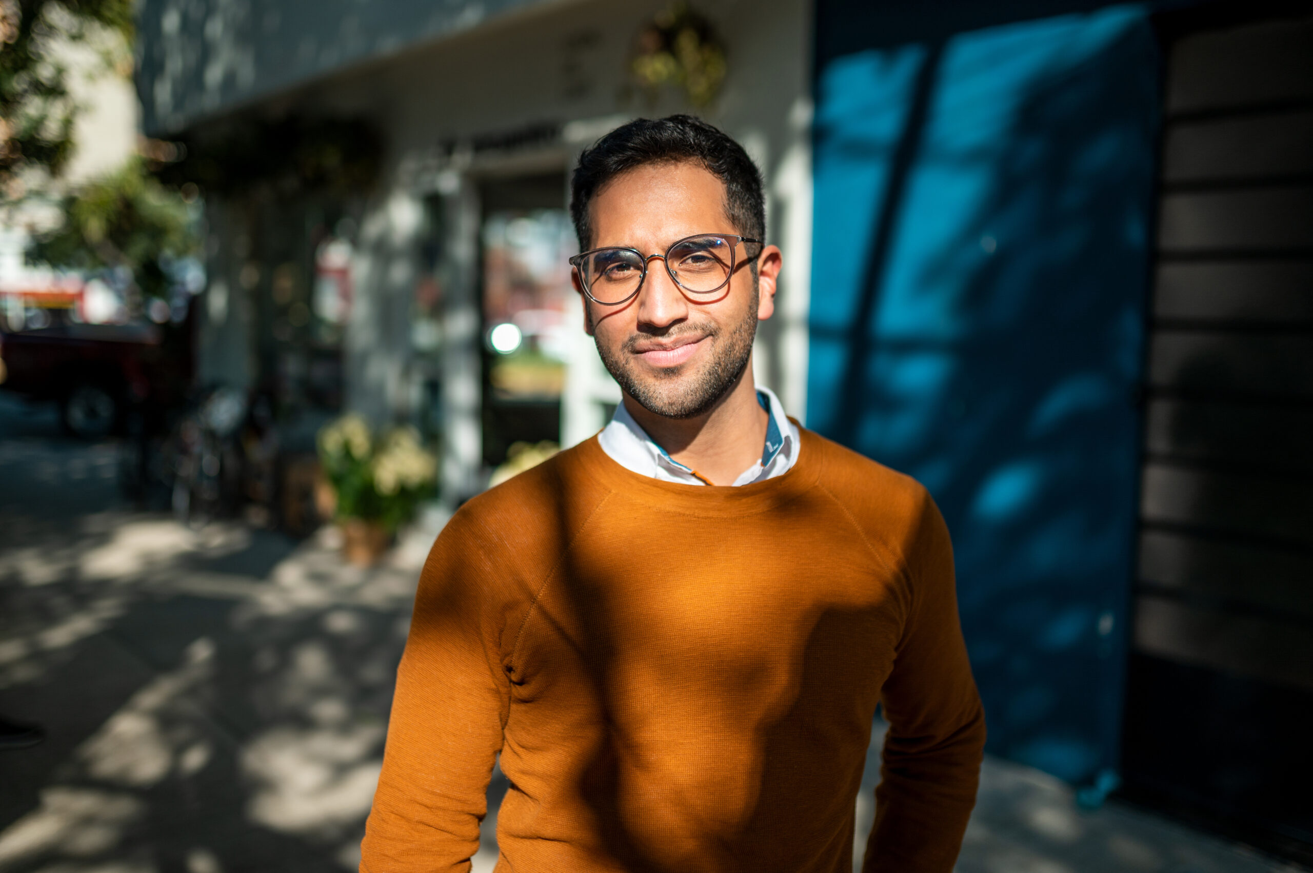Smiling man in an orange sweater and glasses standing outdoors on a city street